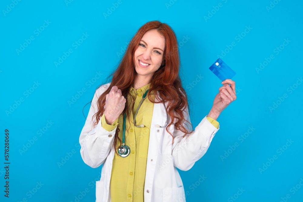 Excited happy positive cheerful smiling young doctor woman wearing medical uniform over blue background hold credit card raise fist in victory Excited happy positive cheerful smiling young doctor woman wearing medical uniform over blue background hold credit card raise fist in victory