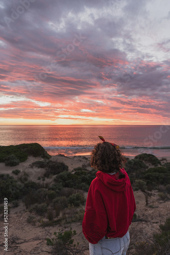 Women with short hair and red hoodie stand up in the sand closed to the beach and looking to the sea and sunset