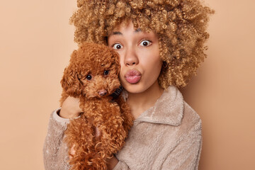 Friendship and domestic pets care. Surprised curly haired woman pouts lips holds poodle puppy near face likes dogs prepare for vet visit stand against brown background. Pet owner poses indoor