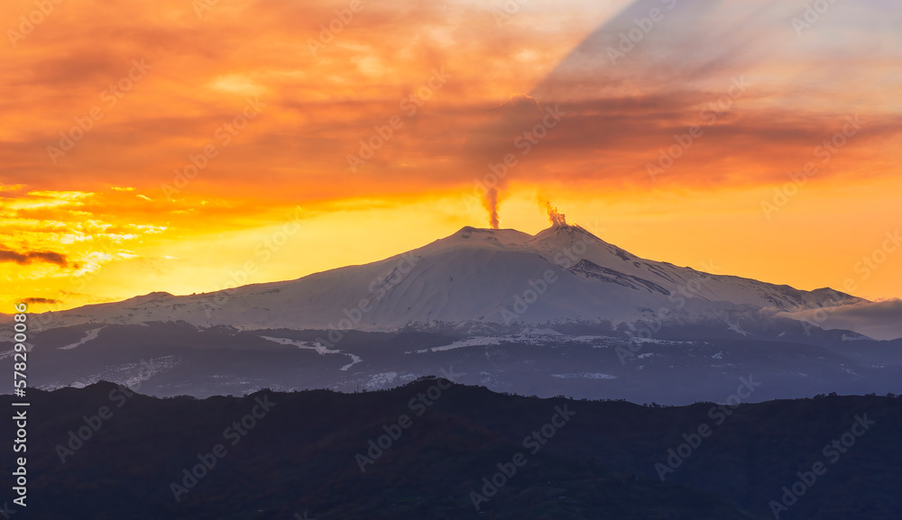 mysterious landscape of great erupting volcano with smoke from craters and snow on slopes in orange light of sunset. eruption of vulcan
