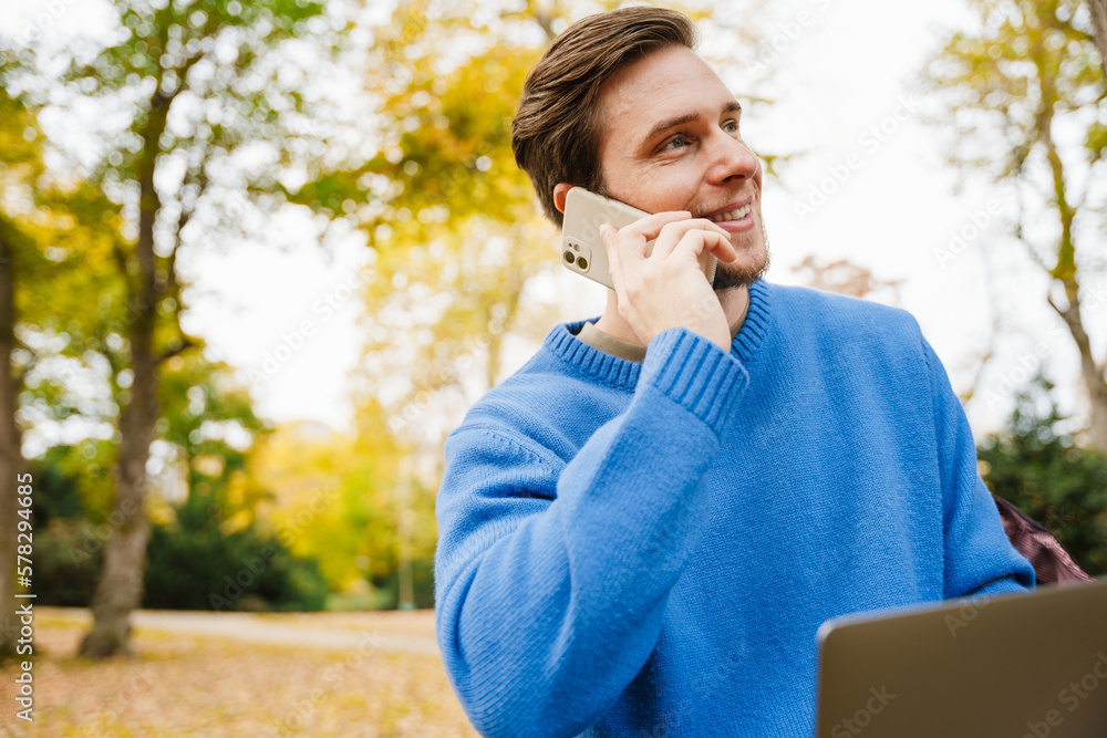 Cheerful man talking on mobile phone while working on laptop in park Cheerful man talking on mobile phone while working on laptop in park