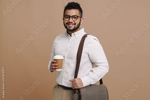 Young indian guy smiling while standing with cup of coffee and bag isolated over beige wall
