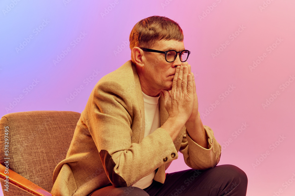Obraz premium Portrait of mature, middle-aged man in jacket sitting on chair with thoughtful face, posing over gradient pink purple background in neon light. Concept of emotions, lifestyle, facial expression, fun