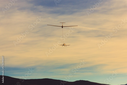 A single-engine aircraft tows a glider against the backdrop of mountains at sunset.