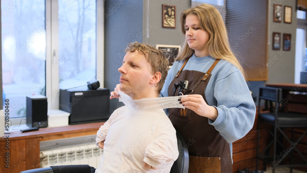 a hairdresser girl prepares a young man with disabilities sitting in a barber chair to cut his hair.