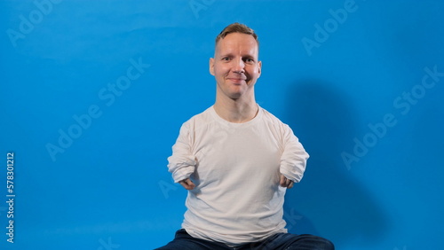 a smiling and cheerful man in a white T-shirt with disabilities sits on an isolated blue background.