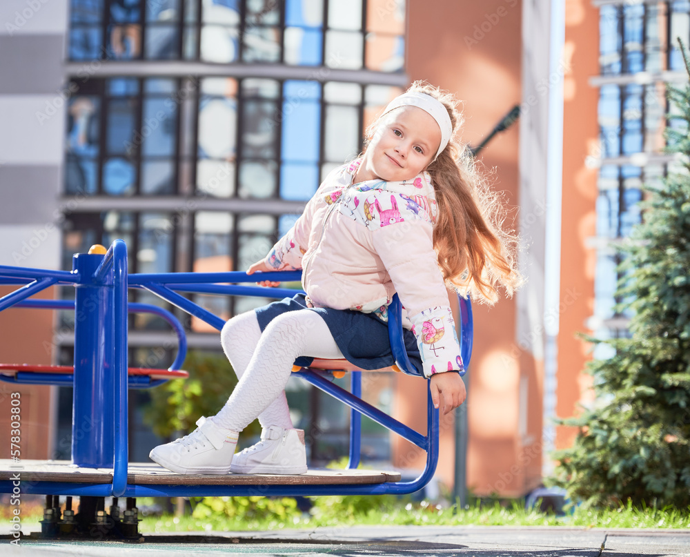 Fototapeta premium Little female child whirling on merry-go-round, having fun at playground. Front view of adorable girl on blue swing, smiling, while revolving outdoors. Modern residential buildings on background.