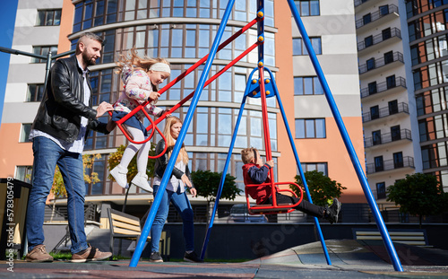 Bearded dad pushing swing with daughter, while mum swinging son next to him in morning. Side view of handsome man, wearing casual, swinging cute daughter in warm autumn day. Concept of parenting.