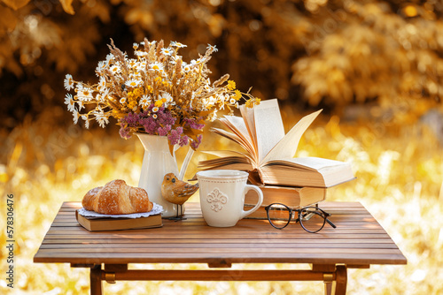 Bouquet of flowers, croissant, cup of tea or coffee, books on table in autumn garden. Rest in garden, reading books, breakfast, vacations in nature concept. Autumn time in garden on backyard