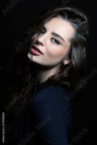 Brunette woman with long wavy hair sensual studio portrait. Model wearing blue blouse and looking at camera with blue eyes and seductive look. Dark studio background. Toned image with blue color