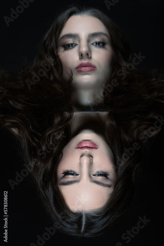 Fototapeta Naklejka Na Ścianę i Meble -  Make-up and fashion concept. Brunette woman with long wavy hair sensual studio portrait. Model looking to reflection of her face in mirror. Dark studio background. Toned image with blue color
