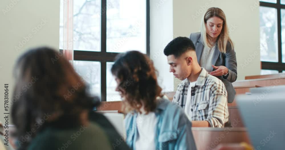 Diverse students sitting at lesson and studying. Inside. Female Caucasian handing out materials for test on background. Woman teacher giving papers with exam. Examination concept. Diverse students sitting at lesson and studying. Inside. Female Caucasian handing out materials for test on background. Woman teacher giving papers with exam. Examination concept.