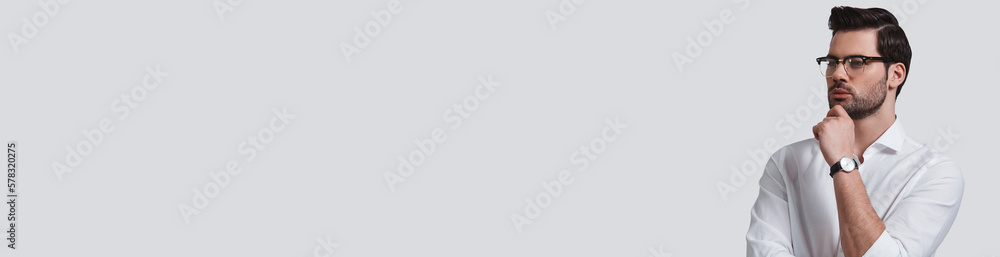 Thoughtful young man in eyeglasses holding hand on chin against grey background Thoughtful young man in eyeglasses holding hand on chin against grey background