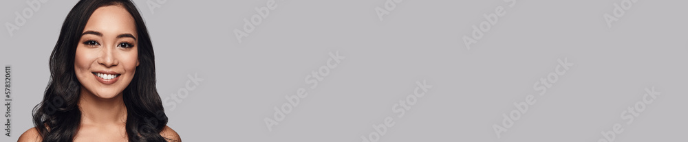Beautiful young woman looking at camera and smiling while standing against grey background Beautiful young woman looking at camera and smiling while standing against grey background