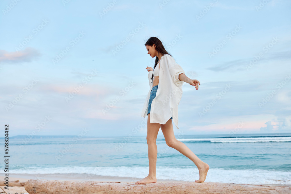 Happy tanned woman in white swimsuit shirt and denim shorts walks the beach on a log on the sand by the ocean with wet hair after swimming, sunset light in Bali Happy tanned woman in white swimsuit shirt and denim shorts walks the beach on a log on the sand by the ocean with wet hair after swimming, sunset light in Bali