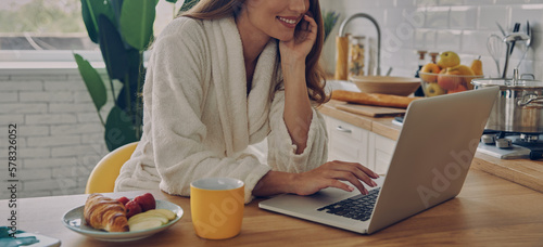Close-up of woman in bathrobe using laptop while having breakfast in the kitchen
