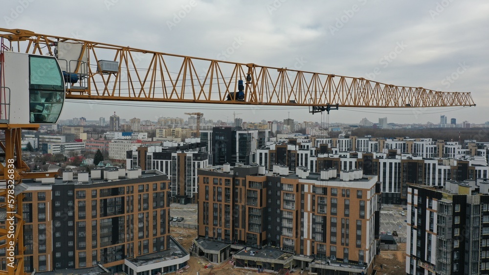 A cab with a crane operator and a yellow construction crane jib towers over a construction site in a residential area. Shooting from a drone at the height of a tower crane against a gray sky