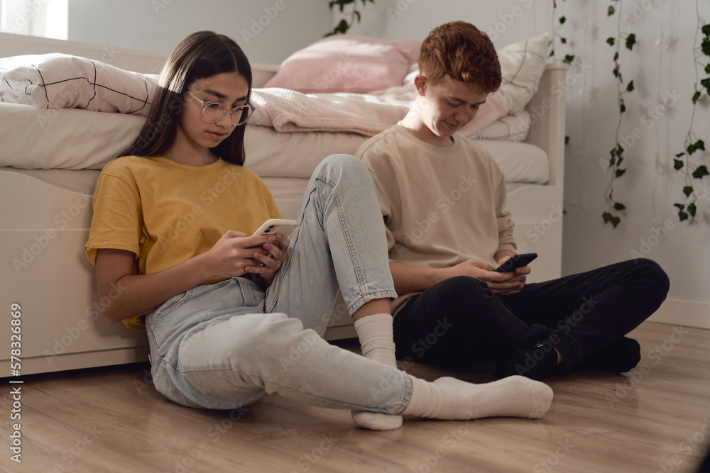 Two caucasian teenage friends sitting on floor in bedroom and browsing phones in silence