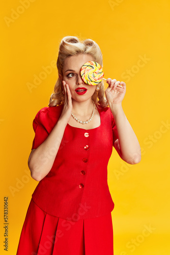 Candy lover. Portrait of beautiful young blonde girl with stylish hairstyle in red suit posing against yellow studio background. Concept of retro fashion, beauty, 50s, 60s. Pin-up style