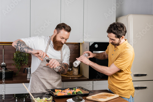 Canvas-taulu Smiling gay couple seasoning chicken fillet and vegetables on baking sheet in kitchen
