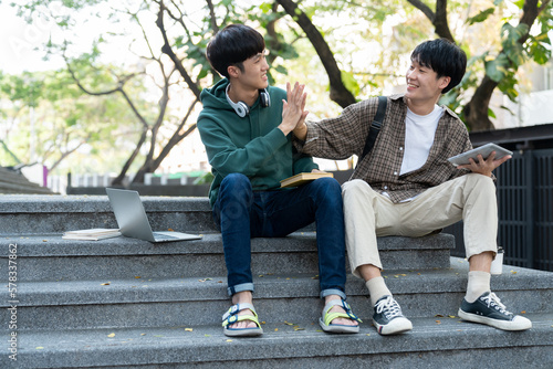 Two Asian male students sitting on the stairs of the university enjoy chatting after school using smartphones and tablets to find information together.