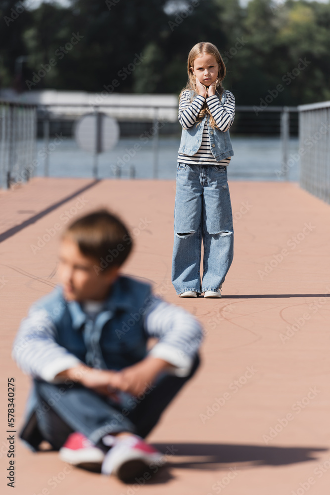 full length of preteen girl in denim outfit standing on river embankment near blurred boy on foreground.