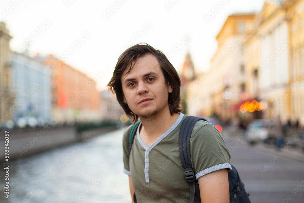 Young male man tourist with medium length hair in old city, river channel. European caucasian white man, looking at camera student, musician, violinist, painter, artist. Lifestyle portrait