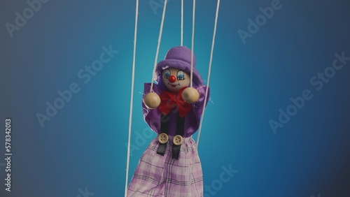 Marionette clown hanging on strings. A rag doll in a purple suit and hat, with a red bow, nose and makeup. Soft doll harlequin funster on a blue studio background. Close up. photo