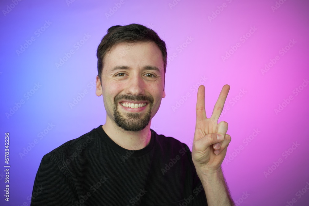 Confident and assertive man showing two fingers in a gesture of victory or peace, set against a captivating backdrop of purple and blue hues. Confident and assertive man showing two fingers in a gesture of victory or peace, set against a captivating backdrop of purple and blue hues.