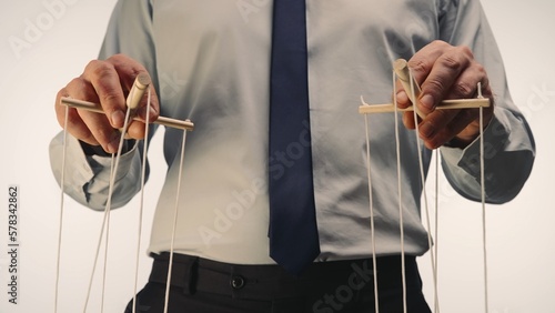 A businessman in a gray shirt and black tie controls a puppet with a wooden manipulator and strings. The puppeteer manipulates the puppet by pulling the ropes with both hands. Close up.