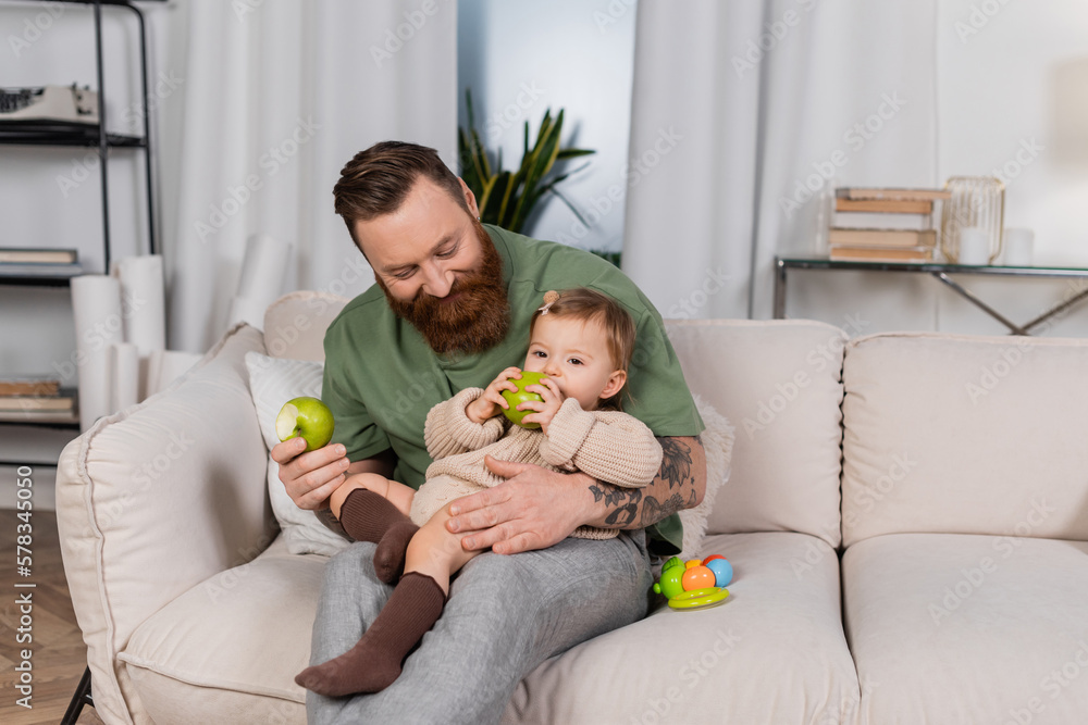 Positive bearded dad holding apple and looking at toddler daughter on couch. Positive bearded dad holding apple and looking at toddler daughter on couch.