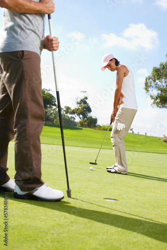 She doesnt let anything distract her. Young woman putting on the golf course with her husband in the background.