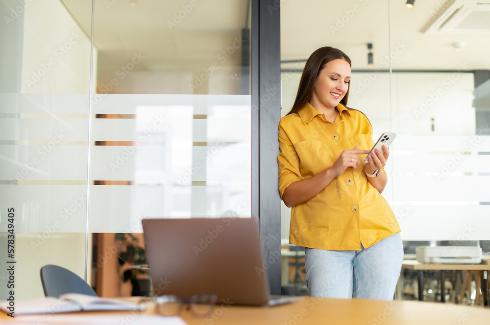 Naklejka premium Cheerful woman texting message at the smartphone while standing at the office meeting room. Business people concept