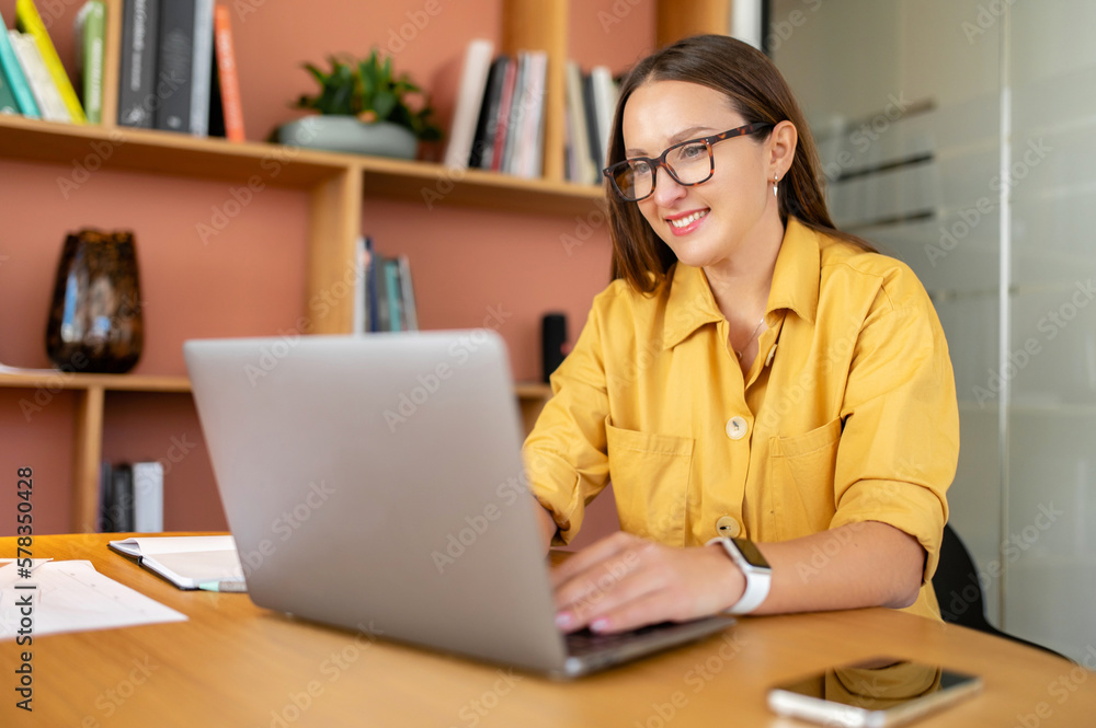 Portrait of young woman in glasses sitting at the desk with laptop, typing an email, messaging colleague, chatting, discussing work moments with pleasant smile Portrait of young woman in glasses sitting at the desk with laptop, typing an email, messaging colleague, chatting, discussing work moments with pleasant smile