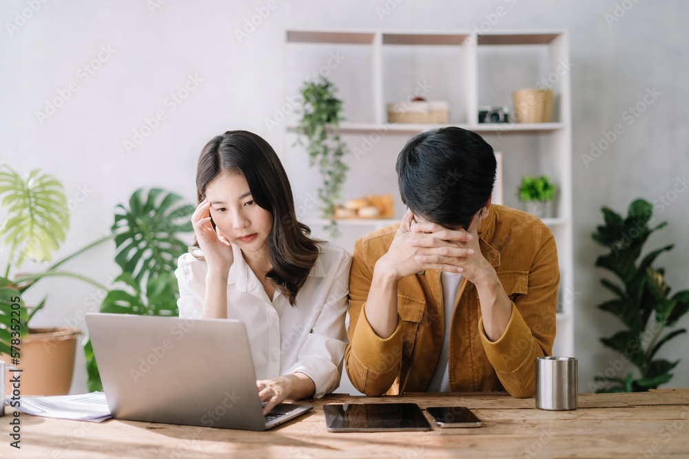 Asian couple sitting at table at home, using laptop to check documents overdue bills, taxes, debt due, bank account balance.lack of money bankruptcy, financial problems concept. Asian couple sitting at table at home, using laptop to check documents overdue bills, taxes, debt due, bank account balance.lack of money bankruptcy, financial problems concept.