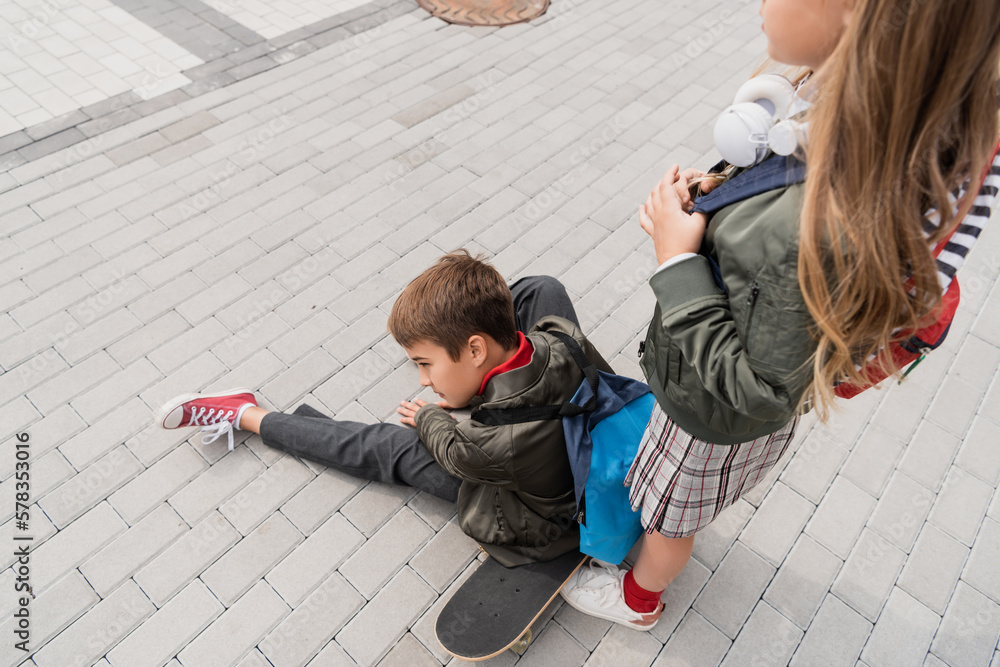 Naklejka premium overhead view of preteen boy in stylish bomber jacket sitting on penny board near girl in wireless headphones.