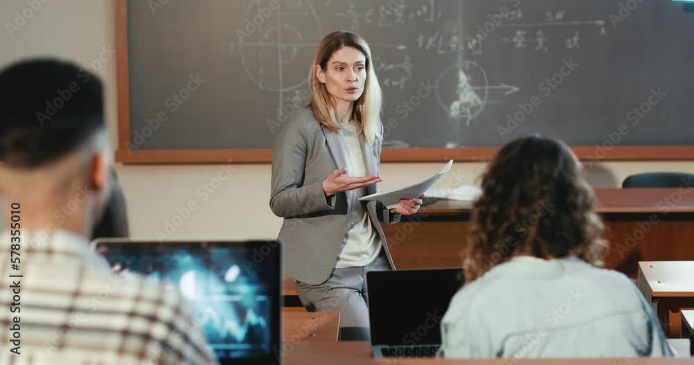 Beautiful Caucasian female professor explaining lection at University in front of students sitting at desks. Education concept. Woman teacher speaking to young adults at college. Geometry math lesson. Beautiful Caucasian female professor explaining lection at University in front of students sitting at desks. Education concept. Woman teacher speaking to young adults at college. Geometry math lesson.