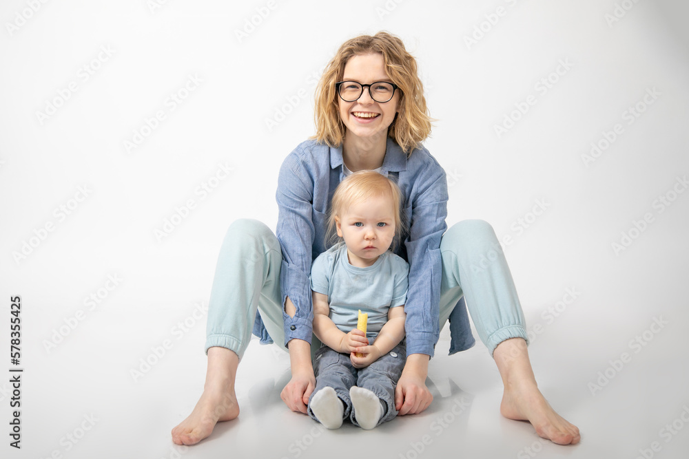 Young charming red-haired woman sits on white background in studio and hugs her little daughter. Care and tenderness Young charming red-haired woman sits on white background in studio and hugs her little daughter. Care and tenderness