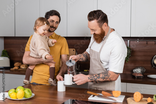 Positive tattooed gay man pouring coffee near partner holding daughter in kitchen.