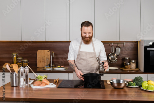 Tattooed man looking at pot on stove while cooking in kitchen.