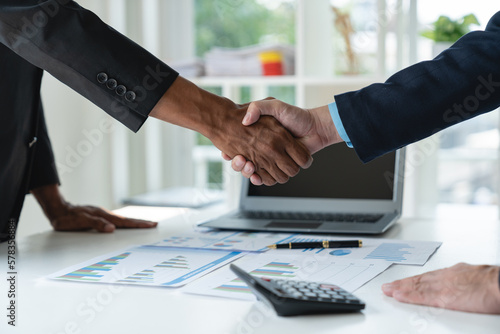 young asian businessman shaking hands with partner after completion and successful financial performance of real estate project Show results from the data graph. Details on laptop at desk.