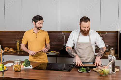 Gay man holding baking paper while partner cooking chicken fillet in kitchen.