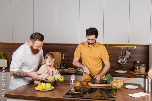 Gay man cooking near partner and toddler daughter in kitchen.