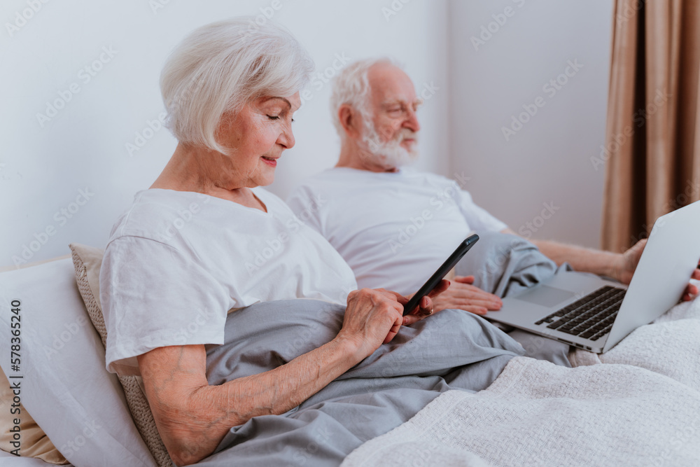 Obraz premium Elderly couple lying in bed at home and using computer laptop and smartphone