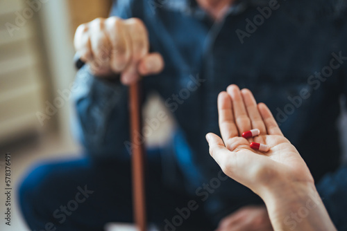 A home healthcare worker helping a senior man in his 70s take his prescription medicine. The patient is in the kitchen, holding a cup of water, looking down as the nurse puts the pills in his hand.