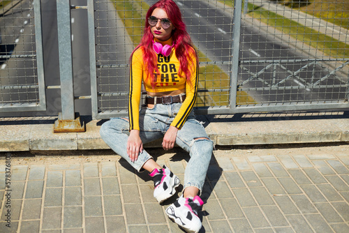 A lady with pink hair in sunglasses, a yellow top and ripped jeans sits on the sidewalk. Vintage retro style clothing from the 2000s in modern fashion. The return of old trends in a new way