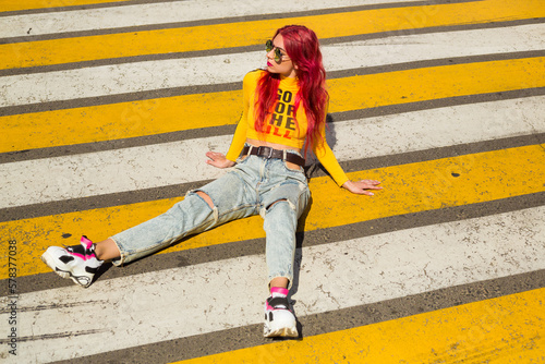 A lady with pink hair in sunglasses, a yellow top and ripped jeans sits on the sidewalk. Vintage retro style clothing from the 2000s in modern fashion. The return of old trends in a new way