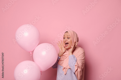 Portrait of amazed happy young Muslim woman in pink hijab and blue casual shirt, celebrating event while standing against pink background with inflatable air balloons. Happy birthday party. Ad space © Taras Grebinets