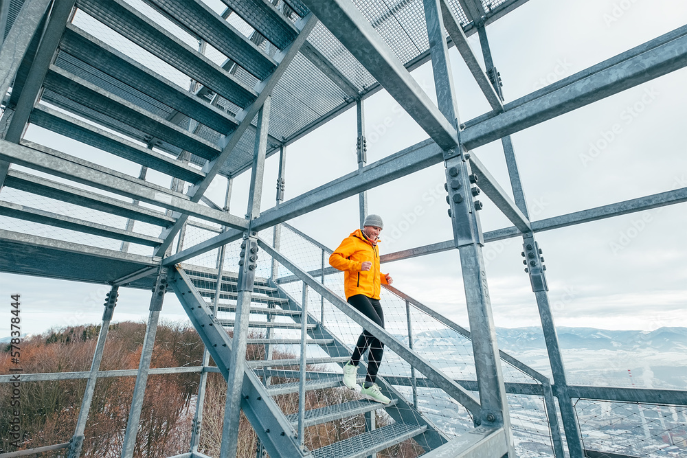 Smiling runner man dressed in bright orange softshell sporty clothes running down by huge steel industrial stairs in cold winter day. People healthy lifestyle concept photo.