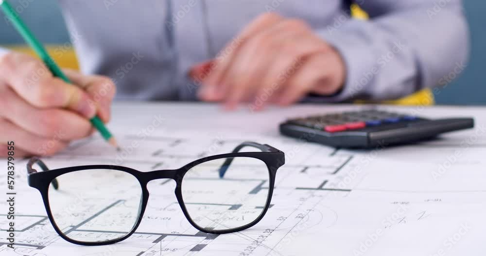 Men's glasses lie on a paper drawing, in the background a man makes notes on paper and calculations on a calculator. Engineer working with exact counts on paper drawing. Men's glasses lie on a paper drawing, in the background a man makes notes on paper and calculations on a calculator. Engineer working with exact counts on paper drawing.